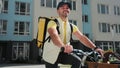 Young courier wearing yellow thermal backpack is making a delivery of the order to the customers by bicycle. Food