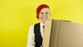 Young crazy man with cardboard box on yellow background in studio. Male courier with bizarre facial expression holds