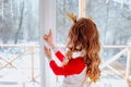 Girl in winter pyjamas looking into the big window for a snowing forest.