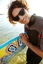 Young curly man in eyeglasses and swimsuit holding surf board