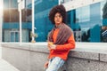 Young curly mixed-race boy drinks coffee on the street against the background of office buildings. Youth culture and fashion. Royalty Free Stock Photo