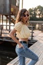 Young curly pretty woman hipster with trendy sunglasses in fashionable jeans and yellow blouse stands on wooden pier by the lake Royalty Free Stock Photo