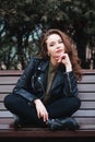 young curly stylish woman wearing black jacket sitting on a bench in city