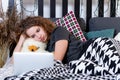 A young curly teen lies in bed caught a teddy bear and looks at the laptop screen