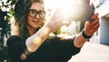 Young curly woman in hat and fashionable glasses makes photo on digital camera of smartphone, standing on city street Royalty Free Stock Photo