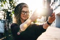 Young curly woman in hat and fashionable glasses makes photo on digital camera of smartphone, standing on city street Royalty Free Stock Photo