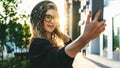 Young curly woman in hat and fashionable glasses makes photo on digital camera of smartphone, standing on city street Royalty Free Stock Photo