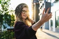 Young curly woman in hat and fashionable glasses makes photo on digital camera of smartphone, standing on city street Royalty Free Stock Photo