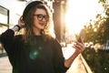 Young curly woman in hat and fashionable glasses makes photo on digital camera of smartphone, standing on city street Royalty Free Stock Photo