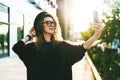 Young curly woman in hat and fashionable glasses makes photo on digital camera of smartphone, standing on city street Royalty Free Stock Photo