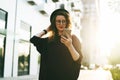 Young curly woman in hat and fashionable glasses makes photo on digital camera of smartphone, standing on city street Royalty Free Stock Photo
