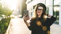 Young curly woman in hat and fashionable glasses makes photo on digital camera of smartphone, standing on city street Royalty Free Stock Photo