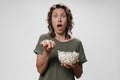 Young curly woman opens eyes and mouth widely eating popcorn, watching a movie or TV Royalty Free Stock Photo