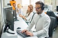 Young customer service operator talking via headset, typing on keyboard and looking at computer monitor in call center Royalty Free Stock Photo