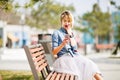 Young cute blond girl with short hair sitting on a wooden bench looks at smartphone and gets surprised wearing denim