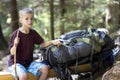 Young cute child boy with stick sitting alone at big tourist backpack on lit by bright summer sun rough wooden bench on green pine Royalty Free Stock Photo