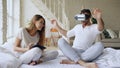 Young cute couple with tablet computer and virtual reality headset playing 360 VR video game while sitting in bed at Royalty Free Stock Photo