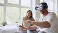 Young cute couple with tablet computer and virtual reality headset playing 360 VR video game while sitting in bed at Royalty Free Stock Photo