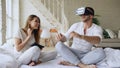 Young cute couple with tablet computer and virtual reality headset playing 360 VR video game while sitting in bed at