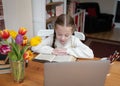 Young cute girl concentrating and working hard reading big old book with laptop and pencils at home on table in white jumper Royalty Free Stock Photo