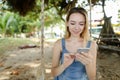 Young cute girl riding swing and using smatrphone, sand and tree in background. Royalty Free Stock Photo