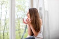 Young and cute lady sitting on the windowsill and looking out th