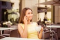 Young cute lady is talking on her smart phone while having coffee in the open air terrace of cafe. She is in elegant outfit, smili Royalty Free Stock Photo