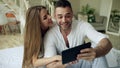 Young cute and loving couple having video chat holding tablet computer and chatting to parents sitting in bed at home Royalty Free Stock Photo