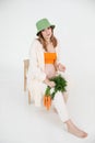 Young cute pregnant woman with long dark hair wear green bucket hat, sitting on chair, holding fresh bunch of carrots. Royalty Free Stock Photo