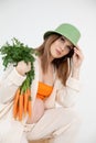 Young cute pregnant woman with long dark hair wear green bucket hat, sitting on chair, holding fresh bunch of carrots. Royalty Free Stock Photo