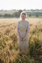 Young pretty girl in yellow dress stands at field of ears in rays of rising sun. grove and village in background Royalty Free Stock Photo