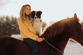 Young happy cute smiling woman with her dog border collie sit on horse in snow field on sunset. yrllow dress Royalty Free Stock Photo