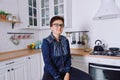 Young cute woman 30+ with short hair in blue jeans and a shirt sitting and relaxing in her clean kitchen Royalty Free Stock Photo