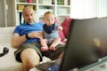 Young dad works remotely from home office with baby. Freelancer man holding his infant while using laptop. Workplace in living Royalty Free Stock Photo