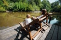 Young dark-haired father and his little son are sitting in recliners on the wooden pier with fishing rods and fishing. Royalty Free Stock Photo