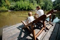 Young dark-haired father and his little son are sitting in recliners on the wooden pier with fishing rods and fishing. Royalty Free Stock Photo