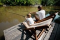 Young dark-haired father and his little son are sitting in recliners on the wooden pier with fishing rods and fishing. Royalty Free Stock Photo