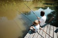 A young dark-haired man and a blond boy dressed in the white t-shirts are sitting with fishing rods on the wooden pier Royalty Free Stock Photo