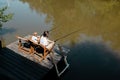 A young dark-haired man and a blond boy are sitting in recliners on the wooden pier with fishing rods and fishing. Royalty Free Stock Photo