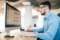 Young dark-haired man in glasess and a blue shirt is working with a computer at his desktop Royalty Free Stock Photo