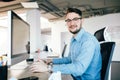 Young dark-haired man in glasses is working with a computer at his desktop in office. He wears blue shirt and smiles to