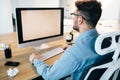 Young dark-haired man is working with a computer at his desktop in office. He wears blue shirt and looks busy. View from Royalty Free Stock Photo