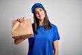 Young delivery woman with blue eyes wearing cap holding paper bag with food with a happy face standing and smiling with a Royalty Free Stock Photo