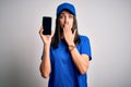 Young delivery woman with blue eyes wearing cap holding smartphone cover mouth with hand shocked with shame for mistake, Royalty Free Stock Photo