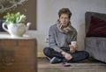 Young depressed looking man with a cold sitting on the floor in his apartment with a thermometer in his mouth and a cup of tea in Royalty Free Stock Photo