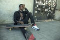 Young depressed and sad black afro American man sitting on grunge street ground corner with skate board feeling depressed