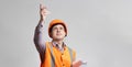 Young design engineer in hard hat and reflective vest on grey background showing with handup, foreman managing