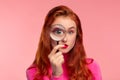 Young detective. Close-up portrait of surprised redhead girl looking at camera through magnifying glass, over pink Royalty Free Stock Photo