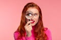 Young detective. Close-up portrait of surprised redhead girl looking at camera through magnifying glass, over pink Royalty Free Stock Photo