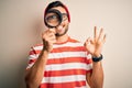 Young detective man looking through magnifying glass over isolated background doing ok sign with fingers, excellent symbol Royalty Free Stock Photo